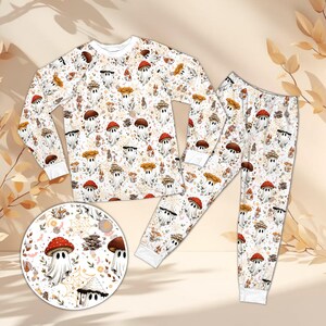 May include: A long-sleeve pajama set featuring a repeating pattern of white ghosts wearing red and brown mushroom caps, interspersed with small spiderwebs, autumn leaves, and various forest fungi on a light cream background.