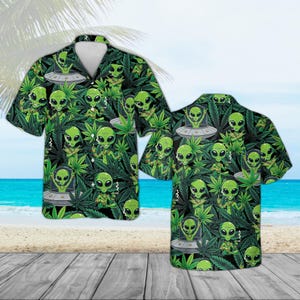 May include: Green short-sleeve button-down shirt with a repeating pattern of green aliens, UFOs, and cannabis leaves. The shirt has a white collar and is displayed on a wooden surface with a beach and ocean background.