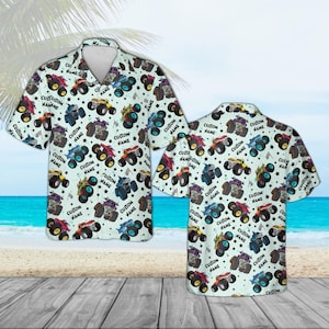 May include: Light blue short-sleeve button-down shirt featuring a repeating pattern of colorful monster trucks. The trucks are in various colors and designs, with the text "CUSTOM NAME" printed on them. The shirt is displayed on a wooden surface.