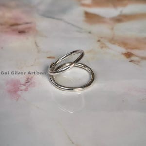 May include: A pair of silver rings, one slightly overlapping the other, on a white and pink marble surface. The rings are simple, circular, and appear to be made of polished metal. The text "Sai Silver Artisan" is visible in the image.