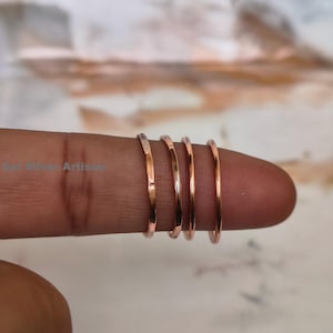 May include: Four thin, rose gold rings are stacked on a finger. The rings have a simple, minimalist design and a polished finish. The rings are a warm, metallic colour and appear to be made of metal. The background is blurred, with neutral tones.