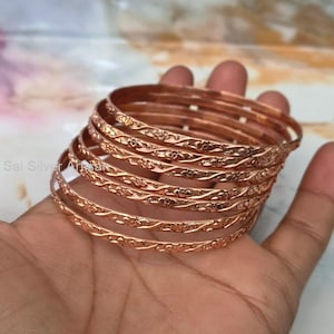 May include: A set of five thin copper-toned bangles featuring a continuous embossed floral pattern. The bracelets are circular with a metallic finish and a repeating vine and flower design that wraps around the entire circumference of each individual band.