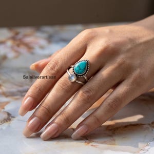 Könnte beinhalten: Ein silberner Ring mit einem tropfenförmigen Türkis und einem kleineren, runden Mondstein. Der Ring befindet sich an einer Hand mit gepflegten Nägeln. Der Shop-Name "Saisilverartisan" ist sichtbar. Der Türkis hat eine Perlenverzierung.