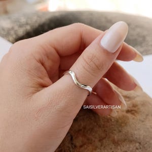 May include: A polished silver ring featuring a smooth, continuous wave-like design that curves gently along the band. The metal has a reflective, high-shine finish. The text SAISILVERARTISAN is visible in the lower right corner of the image.
