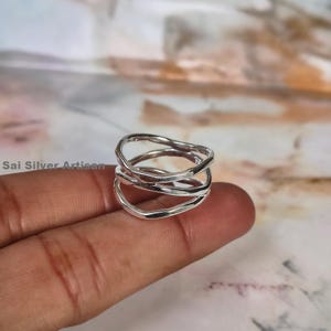 May include: A silver ring with a unique, layered design. The ring features three interconnected, wavy bands, creating an abstract and modern look. The ring is held on a finger, showcasing its size and shape. The ring is from Sai Silver Artisan.