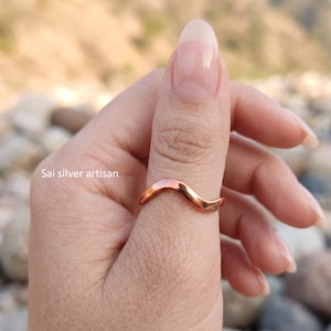 May include: A polished copper ring featuring a smooth, continuous wave-shaped band. The metal has a warm, reddish-brown metallic luster. The text 'Sai silver artisan' is visible on the left side of the image, positioned near the ring.