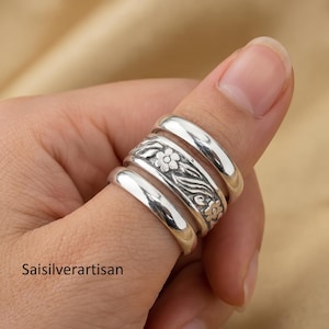 May include: A set of four silver rings on a finger. One ring features a floral design with engraved details. The rings are shiny and appear to be made of sterling silver. The text "saisilverartisan" is visible in the lower left corner.
