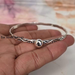 May include: A silver bangle bracelet featuring a polished spherical bead at the center. The band has a wavy, textured design with oxidized detailing along the edges. The text Sai silver artisan is visible on the left side of the metal band.