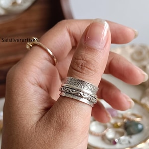 Op de afbeelding: Een brede spinnerring van sterling zilver met drie afzonderlijke banden. De bovenste en onderste banden vertonen ingewikkelde florale en geometrische gravures, terwijl de middelste band een verhoogd, golvend lijnpatroon met kleine stippen heeft.
