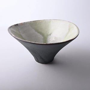 以下が含まれることがあります： 手作りのセラミックボウル。フレアの効いた円錐形。外側は濃いまだら模様のグレーで、内側は淡い緑と白に変化します。縁は繊細で質感のあるエッジです。このユニークなボウルは、盛り付けやディスプレイに適しています。
