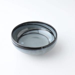 May include: A round, shallow bowl with a glossy finish. The bowl has a gradient of colours, transitioning from a dark black rim to a light blue centre. Thin black lines encircle the bowl's interior. Suitable for serving food. The bowl's diameter is approximately 20 cm.