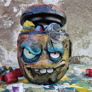 以下が含まれることがあります： 手描きの陶器製容器で、モンスターの顔のデザイン。容器は主に茶色で、青、黄、オレンジのアクセントがあります。黒い蓋は開いており、カラフルなオブジェクトが入っています。ユニークなアート作品。