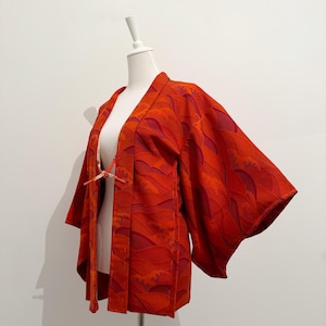 May include: A vibrant orange kimono-style jacket with a wave pattern in shades of red and orange. The jacket features wide sleeves and a tie closure. The garment is displayed on a mannequin against a white background.