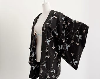 May include: Black kimono-style jacket with a botanical pattern. The jacket features a dark background with a delicate design of white vines and light blue leaves. The sleeves are wide and the jacket is open in the front.