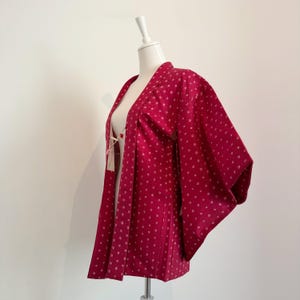 May include: A red kimono-style jacket with a white dotted pattern and floral accents on the shoulders. The jacket is displayed on a mannequin and has wide sleeves and a tie closure. The fabric appears to be lightweight.