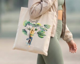 Könnte beinhalten: Eine beige Tragetasche aus Segeltuch mit einer Aquarell-Illustration einer Person in der Baum-Yoga-Pose. Über der Figur ist das Wort Namaste in schwarzer Schreibschrift gedruckt. Das Design zeigt zudem grüne Blätter und Baumzweige.