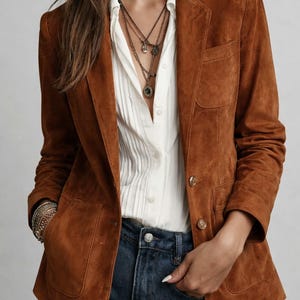 Könnte beinhalten: Ein brauner Wildlederblazer mit zwei Vordertaschen und goldenen Knöpfen. Der Blazer wird über einem weißen Button-Down-Hemd und einer blauen Jeans getragen. Das Outfit wird mit mehrschichtigen Halsketten und Armbändern ergänzt.
