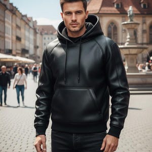 Puede incluir: Una sudadera con capucha de cuero negro con un bolsillo de canguro y una capucha con cordón. La sudadera con capucha se combina con unos vaqueros negros. La persona está de pie en una calle pavimentada con ladrillos con un fondo borroso de edificios y personas.