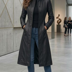 Puede incluir: Un abrigo largo de cuero negro con botones, usado sobre un cuello alto negro y vaqueros azules. El abrigo tiene un corte entallado y bolsillos. Botines negros completan el atuendo. El escenario parece ser una galería de arte.