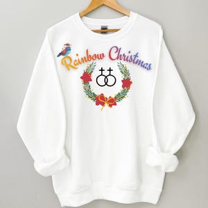 May include: White sweatshirt with the text "Rainbow Christmas" in rainbow colors. A wreath with a red bow and two interlocking circles is in the center. A small bird is on the left side of the text.