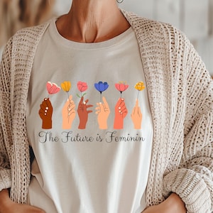 Può includere: T-shirt color crema con una grafica di sette mani che tengono fiori colorati. Le mani sono di diverse tonalità di pelle. Sotto la grafica, il testo recita "The Future is Feminin".