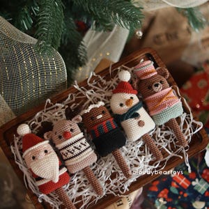 Pode incluir: Uma bandeja de madeira contém cinco picolés de crochê com tema natalino. Os picolés apresentam o Papai Noel, uma rena, um urso e um boneco de neve, cada um com roupas e detalhes únicos. Os picolés são marrons com detalhes brancos, vermelhos e pretos.
