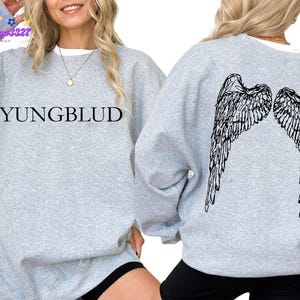 May include: A light gray sweatshirt with the word "YUNGBLUD" printed in black on the front. The back of the sweatshirt features a black graphic of angel wings. The sweatshirt has long sleeves and a crew neck.