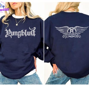 May include: Navy blue sweatshirt with the word "Yungblud" in a gothic font on the front. The back of the sweatshirt features a silver graphic of the band Aerosmith's logo. The sweatshirt has a crew neck and long sleeves.