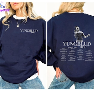 Puede incluir: Sudadera azul marino con ribete blanco. Presenta el texto "YUNGBLUD IDOLS" en la parte delantera. La parte trasera muestra un gráfico de un artista y el texto "YUNGBLUD IDOLS WORLD TOUR 2023".