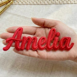 Könnte beinhalten: Ein roter, gestickter Aufnäher mit dem Namen "Amelia" in Schreibschrift. Der Aufnäher wird in der Hand einer Person gehalten, mit einem neutralen Hintergrund. Zwei goldfarbene Stifte befinden sich im Hintergrund.