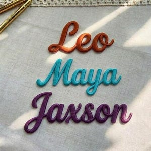 Puede incluir: Parches de nombres bordados en escritura naranja, turquesa y morada. Los nombres "Leo", "Maya" y "Jaxson" se muestran sobre una tela de color claro. Dos bolígrafos dorados están en la esquina superior izquierda.