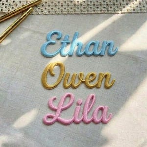 Può includere: Toppe ricamate con nomi in blu, oro e rosa: "Ethan", "Owen" e "Lila". I nomi sono in corsivo. Due spilli color oro sono nell'angolo in alto a sinistra. Le toppe sono su un tessuto chiaro.