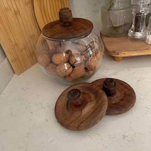Puede incluir: Un tarro de cristal transparente lleno de nueces, coronado con una tapa de madera. Dos tapas de madera adicionales están en primer plano. El tarro y las tapas están sobre una encimera blanca. Las tapas de madera tienen un acabado marrón oscuro.