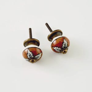 May include: Two round ceramic cabinet knobs featuring a glossy burnt orange finish with white leaf patterns and black outlines. Each knob has a dark antique brass metal base and a threaded metal bolt extending from the back for installation.