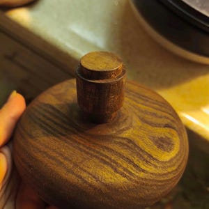 Puede incluir: Una tapa de madera con un pequeño asa cilíndrica. La tapa es redonda con un patrón de vetas arremolinadas en tonos marrones y amarillos. El asa es de un marrón más oscuro.