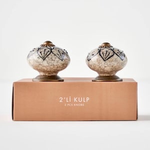 May include: Two oval-shaped ceramic knobs with a cream and brown crackle glaze finish, featuring black painted floral patterns and antique brass-toned metal bases. They are displayed on a tan box labeled 2'Lİ KULP 2 PCS KNOBS.