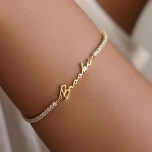 May include: A gold bracelet with the name "Brooke" in cursive. The bracelet has a delicate chain set with small, sparkling stones. The jewellery is shown on a person's arm.