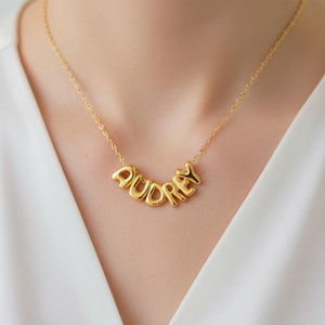 May include: A gold necklace spelling out the name "AUDREY" in bubble letters. The necklace is suspended from a fine chain. The background is a white top.