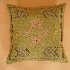 May include: A square, olive green decorative pillow with a woven texture. The pillow features a central geometric design in pink and brown, surrounded by arrow-like patterns and diamond shapes. The edges are trimmed with a dark brown border.
