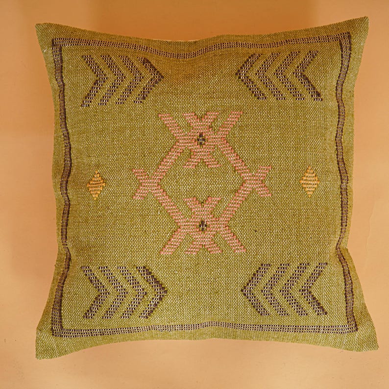 May include: A square, olive green decorative pillow with a woven texture. The pillow features a central geometric design in pink and brown, surrounded by arrow-like patterns and diamond shapes. The edges are trimmed with a dark brown border.