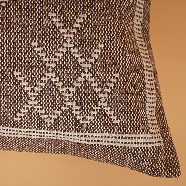 May include: Close-up of a brown and white woven pillow. The pillow features a geometric pattern in white against a brown background. A white border with a double stripe detail frames the edge of the pillow, adding a touch of elegance.