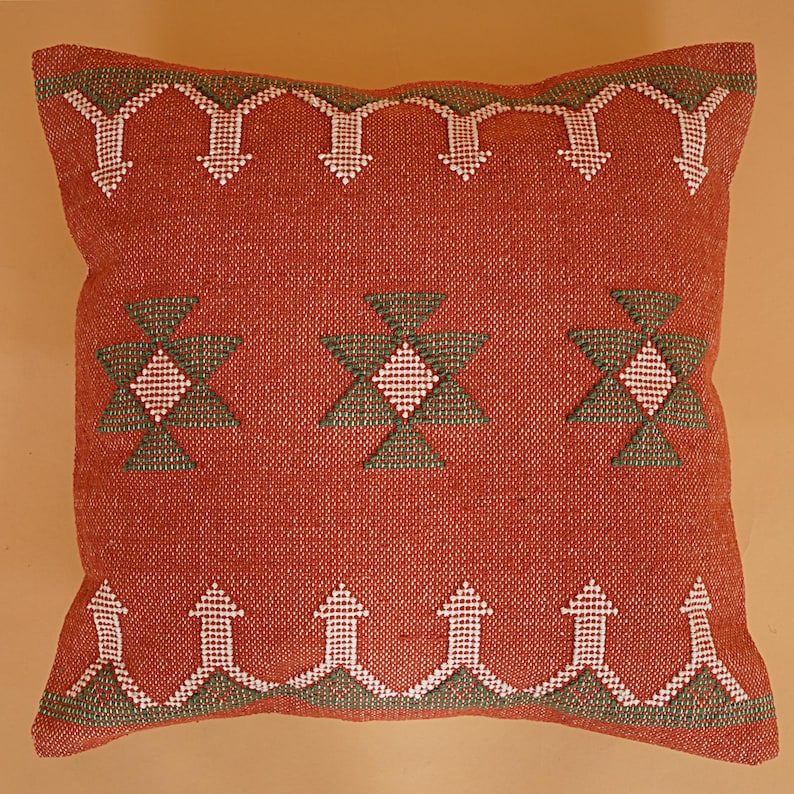 May include: A square, rust-colored decorative pillow with geometric embroidered patterns. The design features white and green arrow and diamond shapes, creating a textured, handcrafted appearance. The pillow is a home decor item.