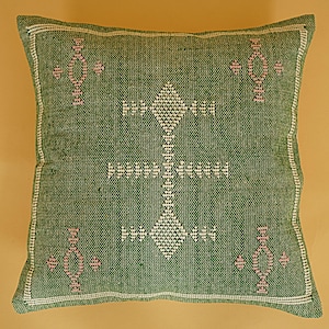 May include: A square green pillow with a white and pink geometric pattern. The pillow has a white border and a textured appearance. The central design is a diamond shape with a vertical line of connected diamonds.