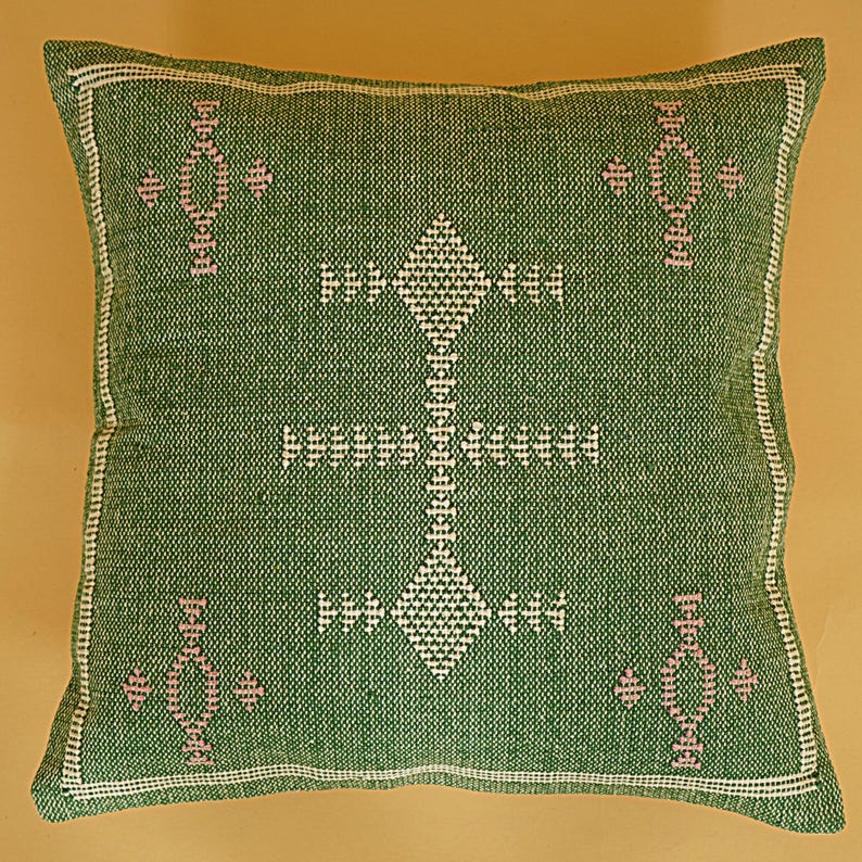 May include: A square green pillow with a white and pink geometric pattern. The pillow has a white border and a textured appearance. The central design is a diamond shape with a vertical line of connected diamonds.