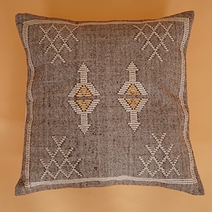 May include: Square brown decorative pillow with white and yellow geometric patterns. The pillow has a textured appearance and a white border. The design includes diamond and cross-like shapes.