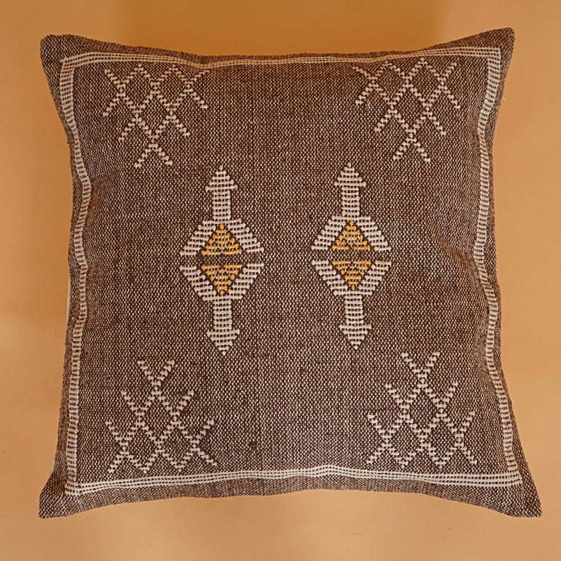 May include: Square brown decorative pillow with white and yellow geometric patterns. The pillow has a textured appearance and a white border. The design includes diamond and cross-like shapes.