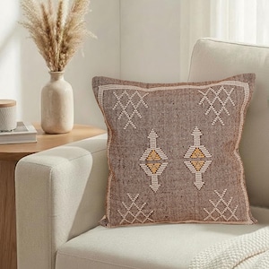 May include: A brown square throw pillow with a geometric pattern in white and yellow. The pillow is on a cream-colored sofa. A vase with dried plants sits on a side table.