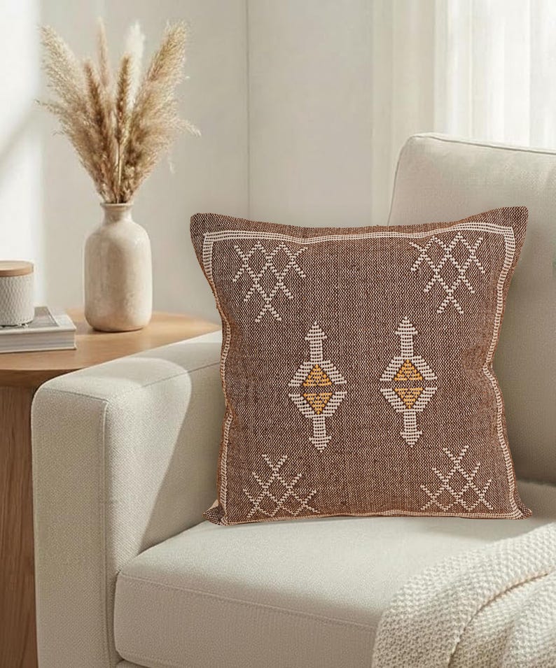 May include: A brown square throw pillow with a geometric pattern in white and yellow. The pillow is on a cream-colored sofa. A vase with dried plants sits on a side table.