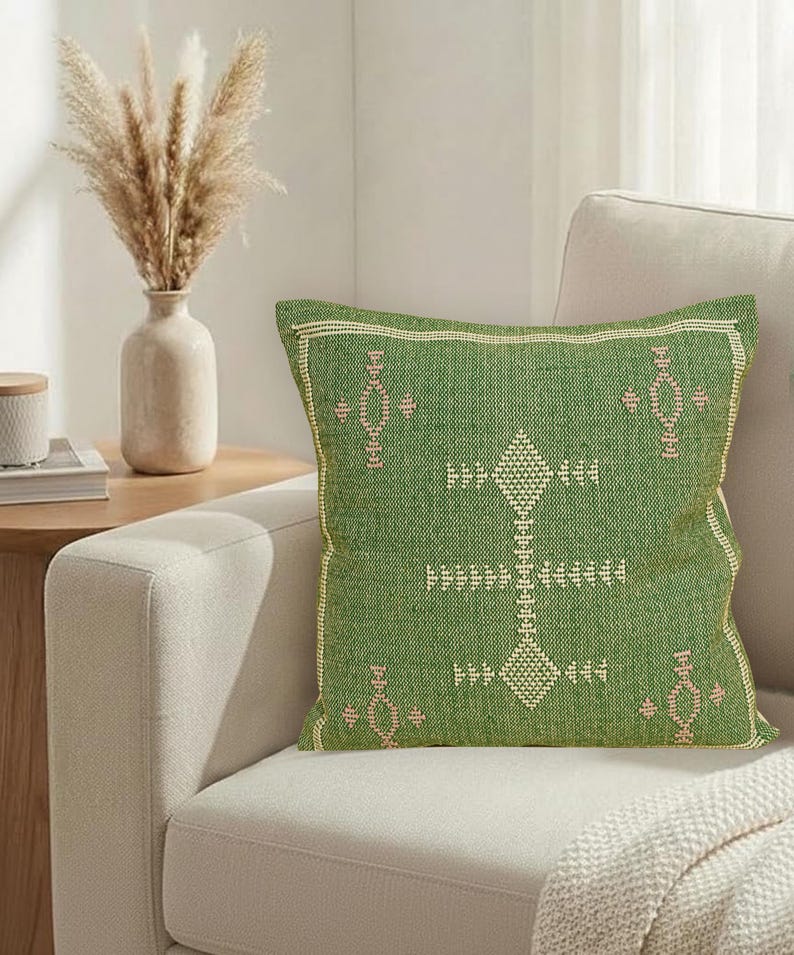 May include: A square green decorative pillow with a geometric pattern in light pink and white. The pillow is on a beige sofa, with a vase of dried flowers and a side table in the background.