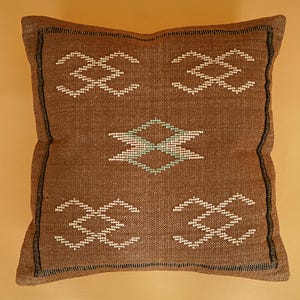 May include: Brown square pillow with a woven texture and geometric patterns. The pillow features white and teal diamond and cross-like designs. The edges are outlined with a black border, creating a decorative accent.